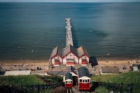 A scenic view overlooking a beach, with a wooden pier extending into the expansive ocean. At the base, a funicular railway leads down to charming red and white timber-framed buildings, characteristic of traditional seaside architecture. People can be seen enjoying the sandy beach and swimming in the sea under a clear blue sky.