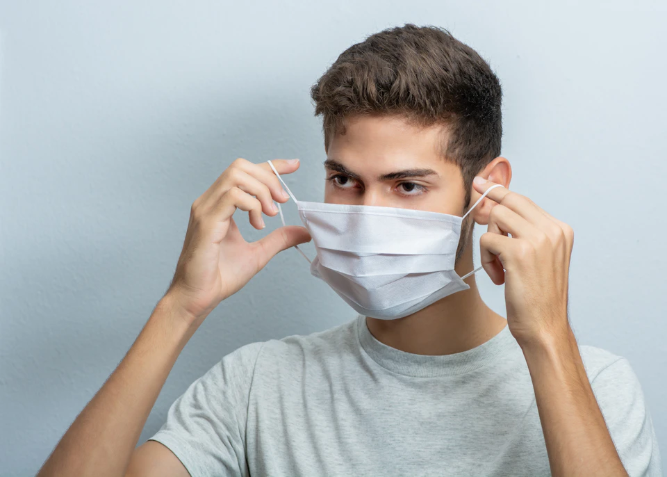 Young man wearing white surgical mask adjusts it with both hands against gray wall backdrop