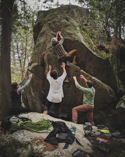 group of people outdoors standing on rocky ground climbning up a big boulder with safety spotters