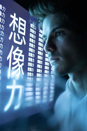 A person intently gazes at a digital screen displaying large, illuminated characters in a dark setting. The characters appear to be in an East Asian script and are repeated multiple times across the screen. The lighting is cool and the scene feels technological and introspective.
