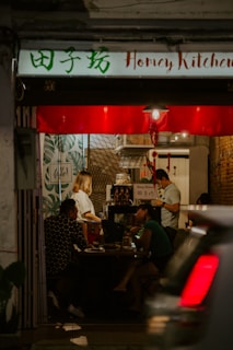A cozy restaurant interior with dim lighting and a small group of people seated at a table. The walls are decorated with leaves and patterns, and a sign reads 'Honey Kitchen'. A person appears to be working behind a counter, and another is engaged in a conversation with the seated guests. A car is partially visible in the foreground, driving past the entrance.