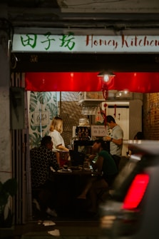 A cozy restaurant interior with dim lighting and a small group of people seated at a table. The walls are decorated with leaves and patterns, and a sign reads 'Honey Kitchen'. A person appears to be working behind a counter, and another is engaged in a conversation with the seated guests. A car is partially visible in the foreground, driving past the entrance.