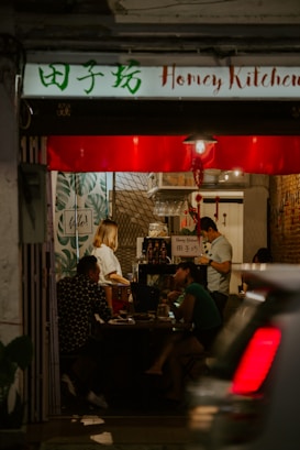 A cozy restaurant interior with dim lighting and a small group of people seated at a table. The walls are decorated with leaves and patterns, and a sign reads 'Honey Kitchen'. A person appears to be working behind a counter, and another is engaged in a conversation with the seated guests. A car is partially visible in the foreground, driving past the entrance.
