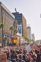 A large crowd of people gathered in an urban area participate in a protest, with many holding signs and raising their fists in the air. The scene takes place on a street lined with tall buildings, including a theater with a neon marquee. Palm trees are visible along the street.