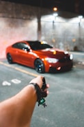 A red sports car is parked in a dimly lit garage. In the foreground, a hand is holding a car key with a small green keychain. The background shows a concrete wall with faint lighting from overhead lamps.