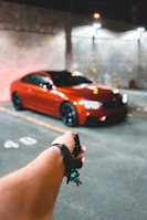 A red sports car is parked in a dimly lit garage. In the foreground, a hand is holding a car key with a small green keychain. The background shows a concrete wall with faint lighting from overhead lamps.