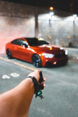 A red sports car is parked in a dimly lit garage. In the foreground, a hand is holding a car key with a small green keychain. The background shows a concrete wall with faint lighting from overhead lamps.