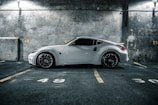 A sleek white sports car is parked in an indoor parking garage with concrete walls. The car is positioned in a parking space marked with the number 49, highlighted by dim overhead lighting. The flooring and walls have a rough, industrial texture, creating a modern and urban atmosphere.