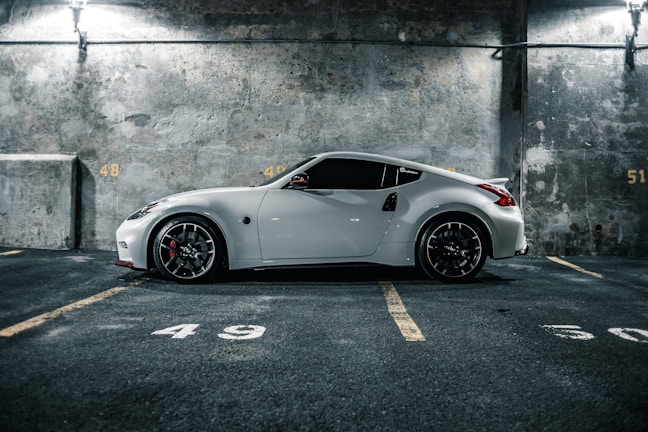 A sleek white sports car is parked in an indoor parking garage with concrete walls. The car is positioned in a parking space marked with the number 49, highlighted by dim overhead lighting. The flooring and walls have a rough, industrial texture, creating a modern and urban atmosphere.