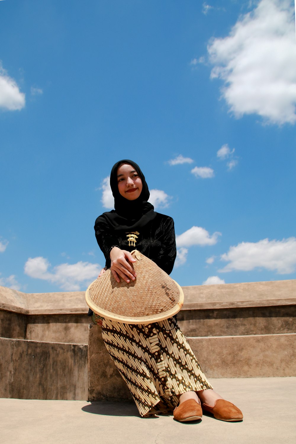 Femme en hijab noir et chemise noire à manches longues assise sur un mur en béton brun pendant la journée