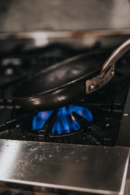 A metal frying pan is positioned on top of a gas stove with a visible blue flame. The pan is slightly tilted, and the kitchen environment appears to be industrial or well-used, with some heat marks and specks visible on the stove surface.