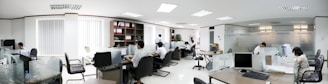 A modern office space with several people working at desks. The room is bright, with white walls and ceiling lights. There are partitions between workstations, and computers are placed on each desk. Shelves with files and plants are visible in the background.