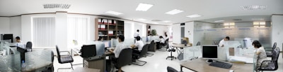 A modern office space with several people working at desks. The room is bright, with white walls and ceiling lights. There are partitions between workstations, and computers are placed on each desk. Shelves with files and plants are visible in the background.