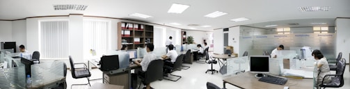 A modern office space with several people working at desks. The room is bright, with white walls and ceiling lights. There are partitions between workstations, and computers are placed on each desk. Shelves with files and plants are visible in the background.