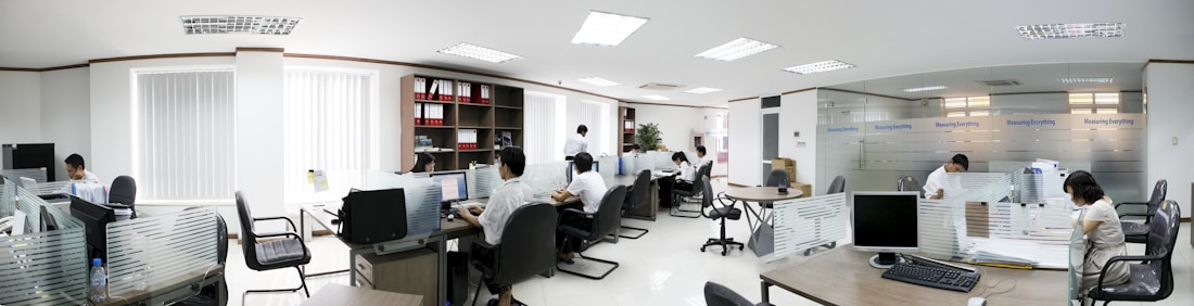 A modern office space with several people working at desks. The room is bright, with white walls and ceiling lights. There are partitions between workstations, and computers are placed on each desk. Shelves with files and plants are visible in the background.