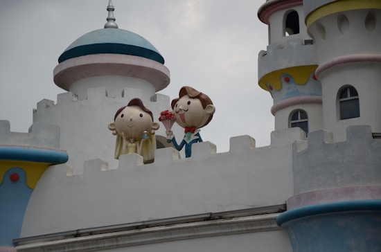 A whimsical castle with cartoon-like figures on the balcony. The characters appear to be a couple, with one figure holding a bouquet of flowers. The architecture includes turrets and decorative elements painted in soft pastels.