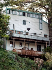 A multi-story wooden house with a modern design is built on a hillside, surrounded by lush greenery and tall trees. The facade features a series of large windows on the upper levels, allowing natural light to flood the interior. The house includes a deck with a wooden railing and a small satellite dish attached. The structure is painted in a light color with green trim, blending harmoniously with the natural environment.