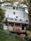 A multi-story wooden house with a modern design is built on a hillside, surrounded by lush greenery and tall trees. The facade features a series of large windows on the upper levels, allowing natural light to flood the interior. The house includes a deck with a wooden railing and a small satellite dish attached. The structure is painted in a light color with green trim, blending harmoniously with the natural environment.