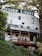 A multi-story wooden house with a modern design is built on a hillside, surrounded by lush greenery and tall trees. The facade features a series of large windows on the upper levels, allowing natural light to flood the interior. The house includes a deck with a wooden railing and a small satellite dish attached. The structure is painted in a light color with green trim, blending harmoniously with the natural environment.