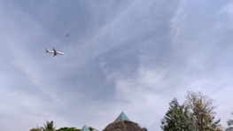 An airplane is flying in a clear sky with a helicopter visible at a distance. Below are lush green trees and foliage, along with a thatched structure crowned by a green triangular roof.