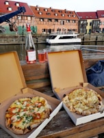 Two boxed pizzas rest on a wooden bench beside a bottle of rosé wine and two cups. The scene overlooks a canal with a docked boat and buildings in the background, featuring red roofs and multiple windows.