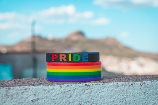 A stack of colorful wristbands, predominantly featuring the rainbow design associated with LGBTQ+ pride. The top band is black with the word 'PRIDE' in multicolored letters. The background is a blurred landscape with a bright blue sky and a distant hill.