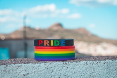 A stack of colorful wristbands, predominantly featuring the rainbow design associated with LGBTQ+ pride. The top band is black with the word 'PRIDE' in multicolored letters. The background is a blurred landscape with a bright blue sky and a distant hill.