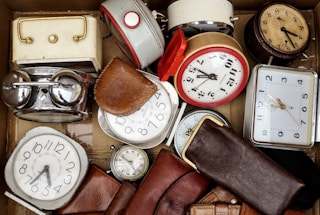 A collection of various vintage alarm clocks and watches are arranged inside a cardboard box. They are surrounded by leather pouches, with some of the clocks having metallic finishes and others with colorful borders.