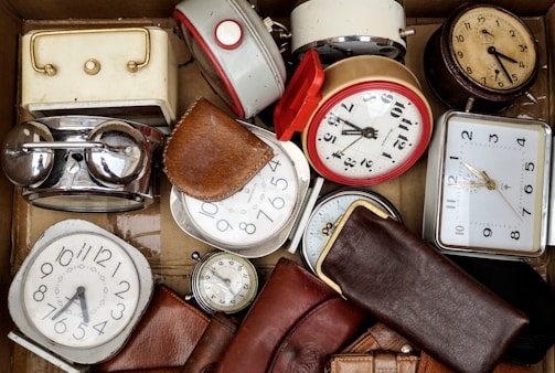 A collection of various vintage alarm clocks and watches are arranged inside a cardboard box. They are surrounded by leather pouches, with some of the clocks having metallic finishes and others with colorful borders.