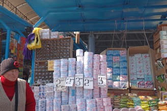 A market stall displays a variety of packaged goods, including rolls of toilet paper with pricing labels. Numerous other packed items, possibly paper goods, are stacked in the background. A person in a red sweater and a black hat featuring a red flower stands in front of the stall.