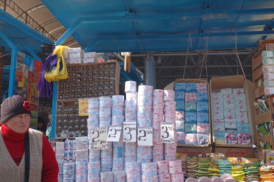A market stall displays a variety of packaged goods, including rolls of toilet paper with pricing labels. Numerous other packed items, possibly paper goods, are stacked in the background. A person in a red sweater and a black hat featuring a red flower stands in front of the stall.
