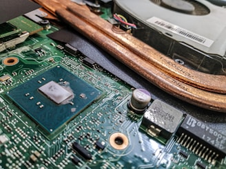 A close-up view of a computer motherboard featuring a large blue central processing unit (CPU) chip surrounded by intricate circuitry and electronic components. Copper heat pipes and a cooling fan are visible in the background, indicating the cooling mechanism for the system.