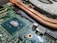 A close-up view of a computer motherboard featuring a large blue central processing unit (CPU) chip surrounded by intricate circuitry and electronic components. Copper heat pipes and a cooling fan are visible in the background, indicating the cooling mechanism for the system.