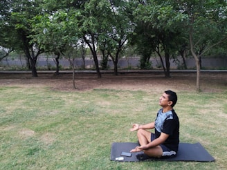 A person is sitting cross-legged on a yoga mat in a grassy area, surrounded by trees. The peaceful setting suggests a meditative or relaxing activity. An electronic device is placed on the mat beside the person.