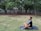 A person is sitting cross-legged on a yoga mat in a grassy area, surrounded by trees. The peaceful setting suggests a meditative or relaxing activity. An electronic device is placed on the mat beside the person.