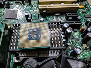 A close-up view of a computer motherboard featuring a prominent central processing unit (CPU) socket with a grid of gold-colored pins. Surrounding the CPU socket are heat sinks, capacitors, and various connectors, with intricate circuit patterns and components.