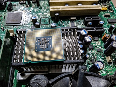 A close-up view of a computer motherboard featuring a prominent central processing unit (CPU) socket with a grid of gold-colored pins. Surrounding the CPU socket are heat sinks, capacitors, and various connectors, with intricate circuit patterns and components.
