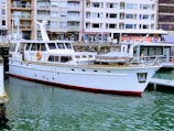 A white yacht docked in a marina with buildings in the background. The water is calm, and the yacht has a red trim at the waterline. Various windows and lifebuoys are visible on the yacht.