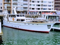 A white yacht docked in a marina with buildings in the background. The water is calm, and the yacht has a red trim at the waterline. Various windows and lifebuoys are visible on the yacht.