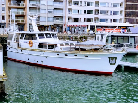 A white yacht docked in a marina with buildings in the background. The water is calm, and the yacht has a red trim at the waterline. Various windows and lifebuoys are visible on the yacht.