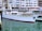 A white yacht docked in a marina with buildings in the background. The water is calm, and the yacht has a red trim at the waterline. Various windows and lifebuoys are visible on the yacht.