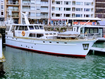 A white yacht docked in a marina with buildings in the background. The water is calm, and the yacht has a red trim at the waterline. Various windows and lifebuoys are visible on the yacht.