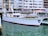 A white yacht docked in a marina with buildings in the background. The water is calm, and the yacht has a red trim at the waterline. Various windows and lifebuoys are visible on the yacht.