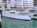 A white yacht docked in a marina with buildings in the background. The water is calm, and the yacht has a red trim at the waterline. Various windows and lifebuoys are visible on the yacht.