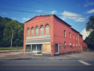 A red brick building with arched windows and a boarded-up entrance sits by a road. The sign reads 'Olga Coal Company' against a backdrop of lush green trees and a clear blue sky.