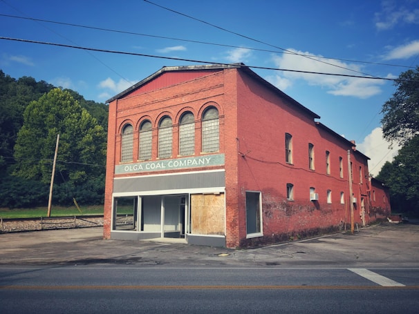 A red brick building with arched windows and a boarded-up entrance sits by a road. The sign reads 'Olga Coal Company' against a backdrop of lush green trees and a clear blue sky.