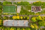 An aerial view reveals a rectangular sports field with a bright green surface, surrounded by a residential area. To the right of the field is a playground with colorful equipment. Numerous trees with lush green and yellow foliage frame the areas, and several cars are parked on the street adjacent to the building and sports field.
