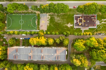 An aerial view reveals a rectangular sports field with a bright green surface, surrounded by a residential area. To the right of the field is a playground with colorful equipment. Numerous trees with lush green and yellow foliage frame the areas, and several cars are parked on the street adjacent to the building and sports field.