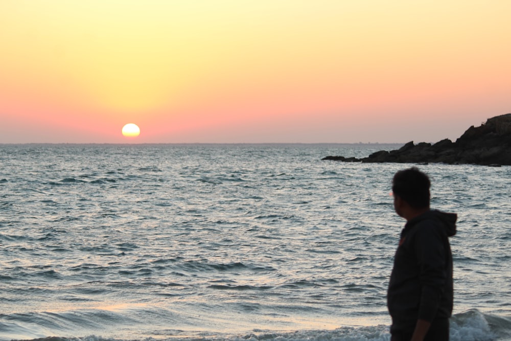 man in black jacket standing on seashore during sunset