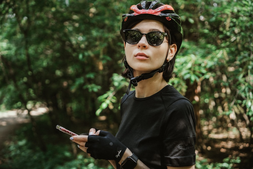 Donna in camicia girocollo nera che indossa occhiali da sole neri e berretto rosso e nero che tiene lo smartphone